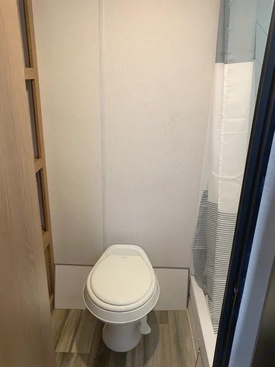 This RV rental bathroom features a clean white toilet, wood-look floor, and a gray/white shower curtain for a comfortable trip.. Avenger LT 2022