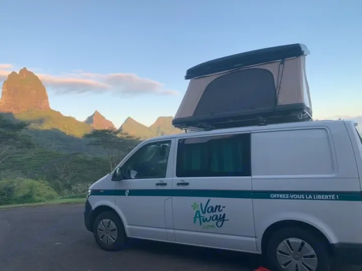 This white Van-Away rv rental campervan with a pop-up roof tent is parked before stunning, sunlit mountains.. Volkswagen Transporter T6.1 2021