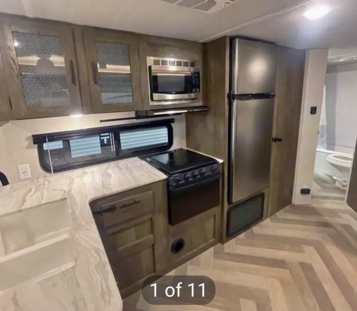 Inside an RV rental: a modern kitchen with stainless steel appliances (fridge, microwave, stove), sink, and wood-look floor. Bathroom visible.. Forest River Wildwood X-Lite 2020