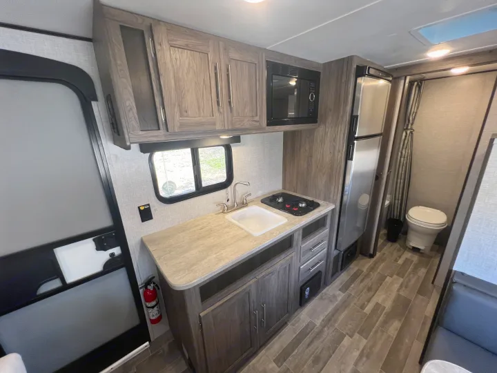 Clean RV rental kitchen: sink, stove, microwave, fridge, and modern wood-grain cabinets. Toilet visible to the right.. Crossroads Zinger Lite 2022
