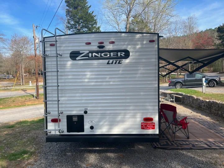 Rear of a white Zinger Lite travel trailer, an RV rental, parked at a gravel campsite with an extended awning and a truck nearby.. Crossroads Zinger Lite 2022