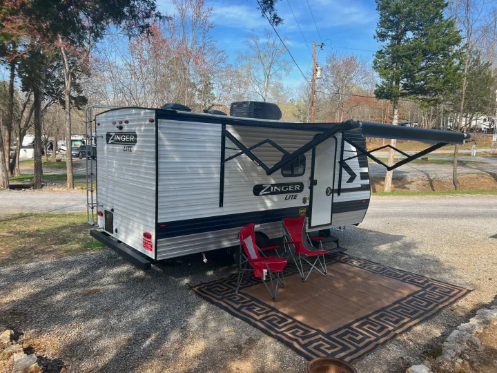 A Zinger Lite travel trailer RV rental, parked on a gravel campsite. Awning extended, with outdoor rug and chairs for relaxation.. Crossroads Zinger Lite 2022