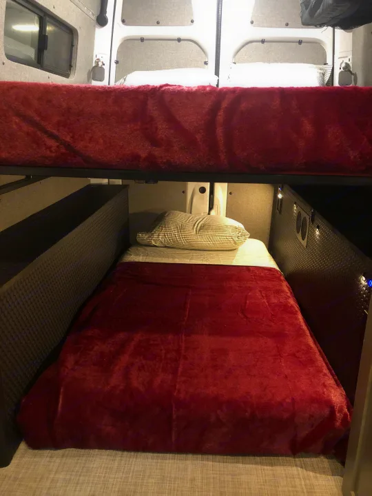 This RV rental features cozy bunk beds. The lower bunk has a striped pillow and red blanket, with textured walls and small lights.. Freightliner Sprinter 2012