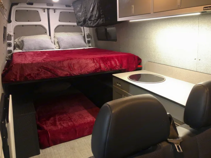Inside an RV rental: a raised bed with red bedding, a lower bunk, and a compact kitchen counter with sink.. Freightliner Sprinter 2012