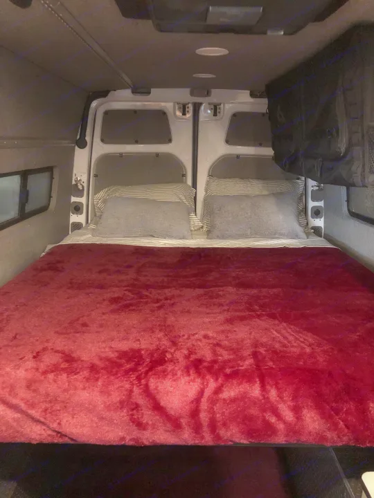 Cozy RV rental van bed with a plush red blanket and pillows, positioned in the vehicle's rear sleeping area.. Freightliner Sprinter 2012