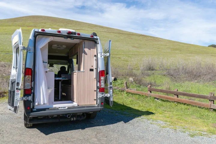 RV rental camper van with open rear doors, revealing its functional interior with a toilet, kitchen, and person inside. Parked by a green hill.. Hymer Sunlight Van 1 2018