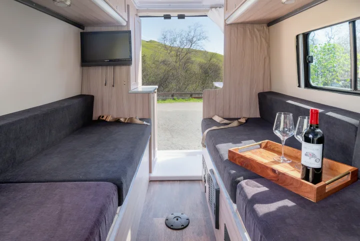 This rv rental offers a comfortable interior with dark upholstered seating, TV, wine setup, and scenic outdoor views.. Hymer Sunlight Van 1 2018