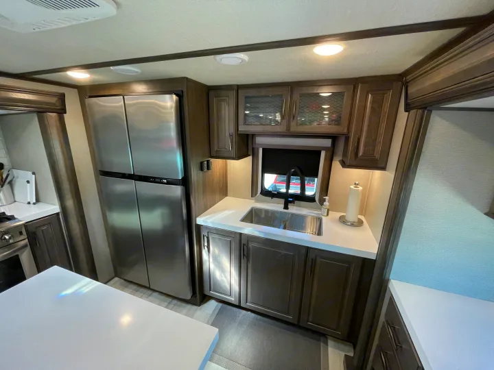 Modern rv rental kitchen with stainless steel appliances, white countertops, dark wood cabinets, and a sink. Brightly lit for comfortable use.. Grand Design Solitude 2022