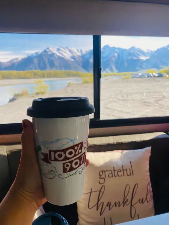 A hand holds a "100% 907" mug inside an RV rental, admiring snow-capped mountains & a river. Grateful & thankful travels!. Winnebago Minnie Winnie 2020