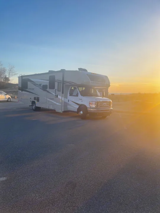A large white RV rental, a Coachmen motorhome, parked during a golden sunset, suggesting a scenic travel stop.. Coachmen Leprechaun 2023