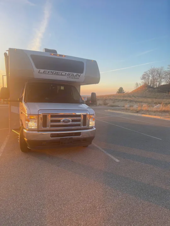 Golden light illuminates this Coachmen Leprechaun RV rental parked at a scenic lookout with sandy hills.. Coachmen Leprechaun 2023