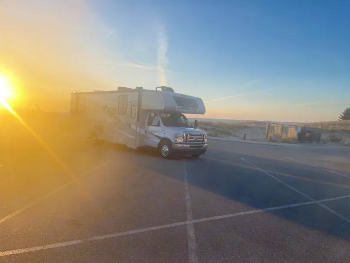 Class C RV rental at sunset, parked by sandy dunes near the water. Golden light illuminates the scene.. Coachmen Leprechaun 2023