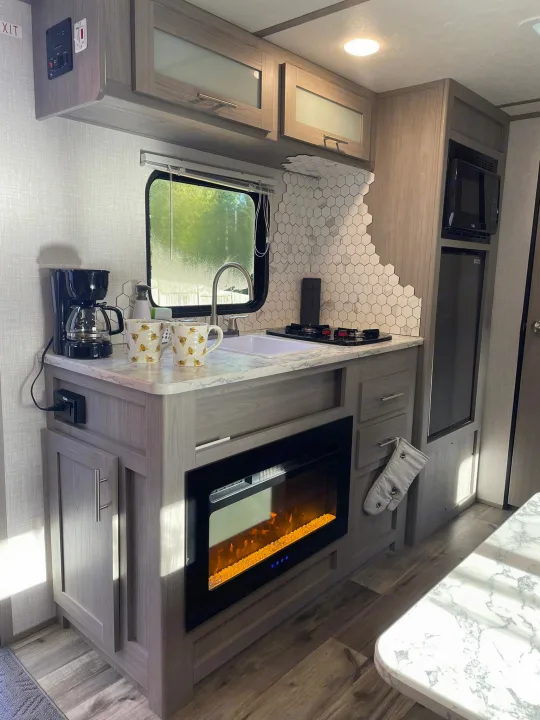 Modern RV rental kitchen with compact design. Features include a sink, stove, microwave, and electric fireplace for cozy stays.. Dutchmen RV Coleman Lantern LT Series 17B 2023