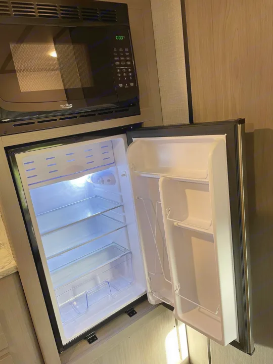 RV rental kitchen: A black High Pointe microwave sits above an open, empty mini-fridge with bright interior lighting.. Dutchmen RV Coleman Lantern LT Series 17B 2023