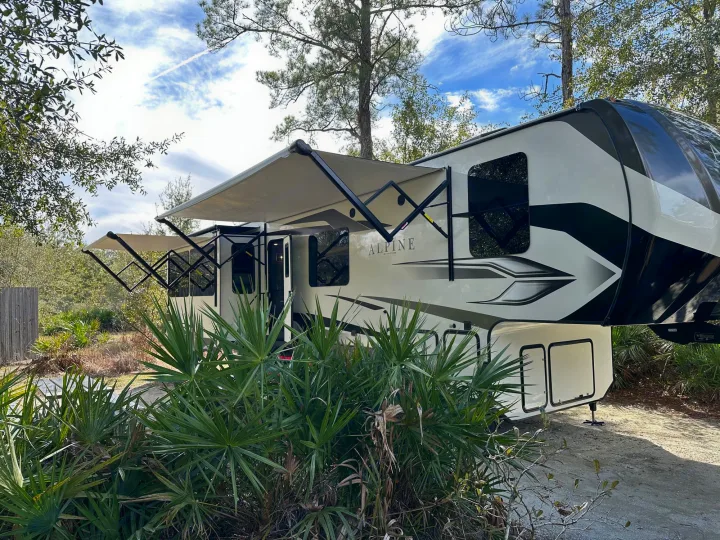 Large, modern RV rental with awnings extended, parked in a sunny camp spot surrounded by trees and palmetto plants.. Keystone RV Alpine 2022