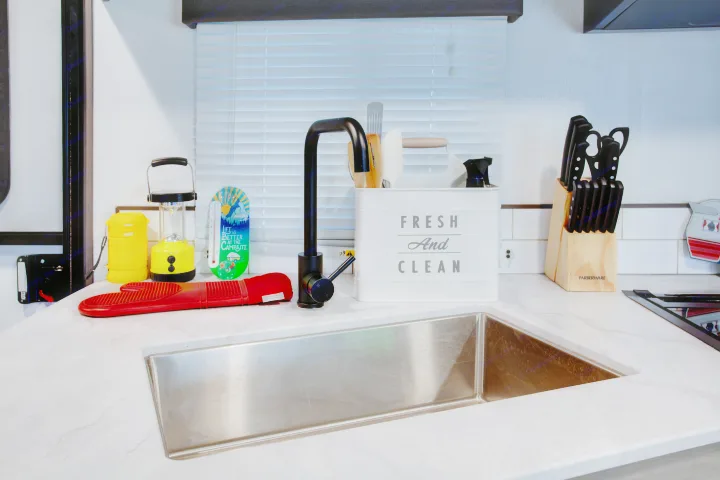 A neat RV rental kitchen workspace: white counters, a stainless steel sink, black faucet, and organized camping-themed essentials.. Cruiser Rv Corp Shadow Cruiser 2021