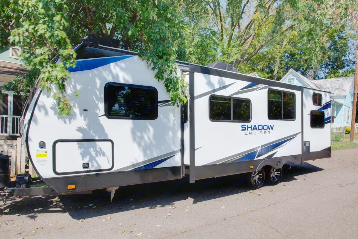A white & blue Shadow Cruiser travel trailer, perfect for an RV rental, is parked with its side extended under tree shade.. Cruiser Rv Corp Shadow Cruiser 2021