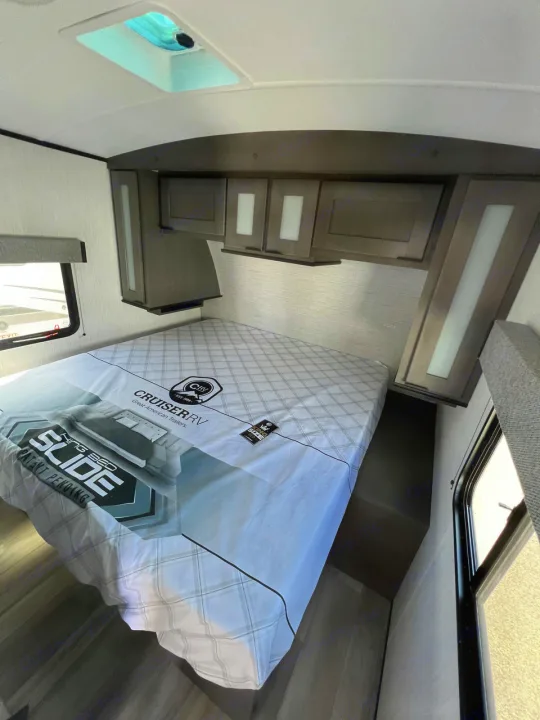 RV rental bedroom with a large bed featuring a 'Cruiser RV' cover. Dark wood overhead cabinets and a ceiling skylight are visible.. Cruiser Rv Corp MPG 2700TH 2022