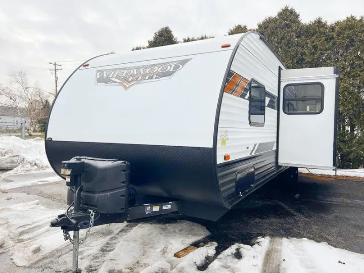 This white Wildwood Lite travel trailer, with its side door open, is parked in a snowy lot, perfect for your next RV rental.. Forest River Wildwood X-Lite 2020