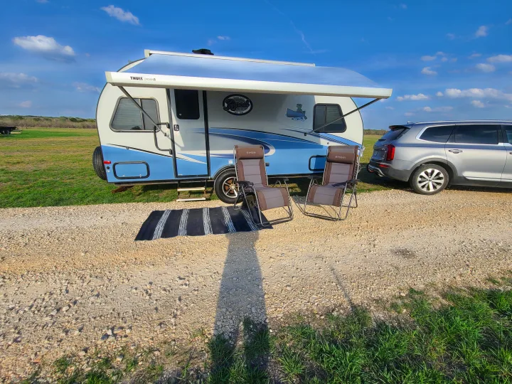 Blue and white travel trailer RV rental parked on gravel, with extended awning, two chairs, and a mat. Ready for camping!. Forest River R-Pod 2018