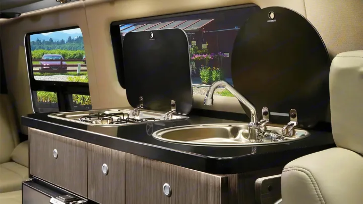 Compact Dometic kitchen inside an rv rental, featuring a stove, sink, and storage units, with a serene outdoor vista.. Airstream Interstate 2021