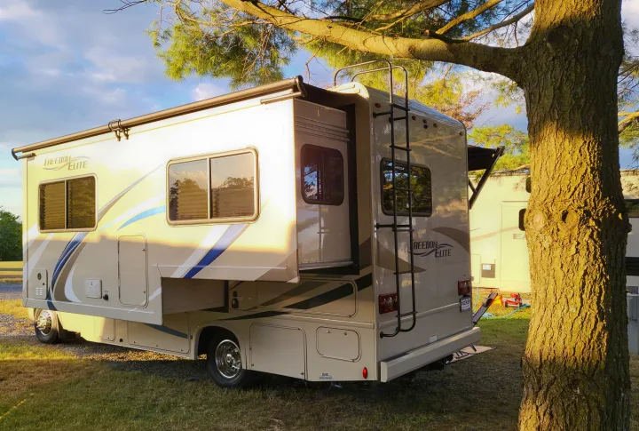 A Freedom Elite RV rental with slide-out extended, parked at a sunny campsite beside a large tree.. Thor Freedom Elite 2018