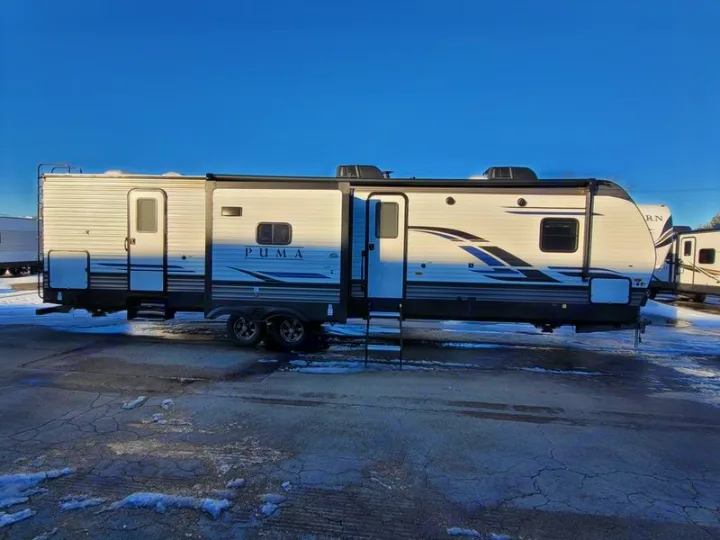 This long white Puma travel trailer, ideal for an RV rental, is seen on a snowy lot. Features extendable sections & multiple doors.. Forest River Palomino Puma 2022