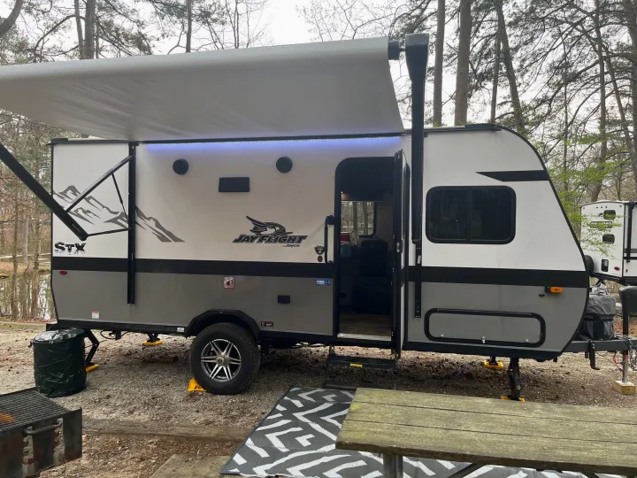 A modern Jayco travel trailer with an extended lighted awning, set up in a wooded campsite. Ideal for an RV rental.. Jayco Jay Flight SLX 2022