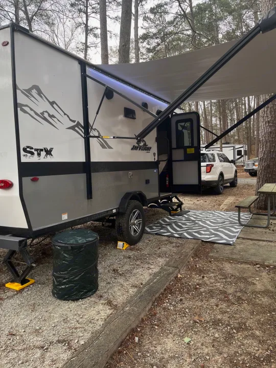 A cozy RV rental campsite featuring a travel trailer with a lit awning, outdoor mat, and a white SUV nearby.. Jayco Jay Flight SLX 2022