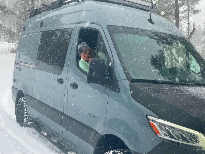 A gray Entegra camper van, perfect for an RV rental adventure, travels through a snowy forest with a happy person and dog inside.. Mercedes-Benz Entegra Launch 2023