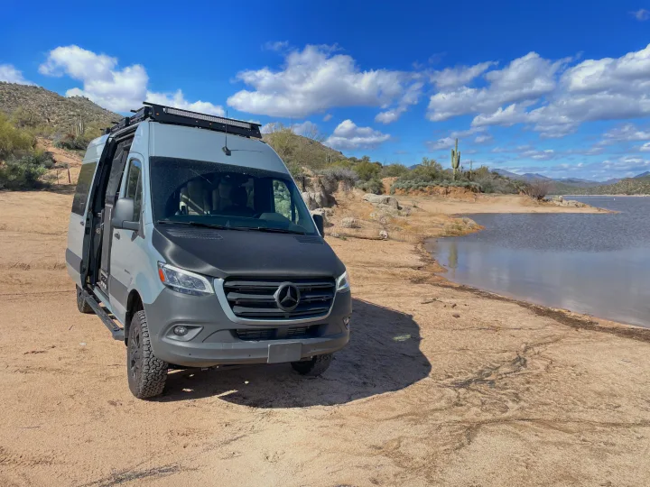 Explore scenic desert landscapes with an RV rental. This Sprinter van is parked by a tranquil lake with mountains and cacti.. Mercedes-Benz Entegra Launch 2023