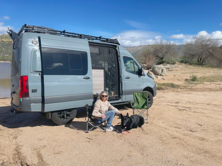 Serene RV rental scene: Woman and dog relax by a gray camper van overlooking a lake. Perfect for outdoor travel!. Mercedes-Benz Entegra Launch 2023