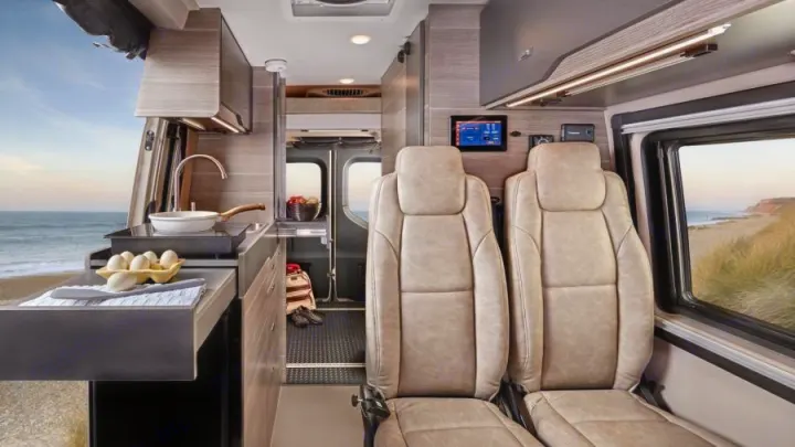 This RV rental showcases a modern interior: kitchen with stove/sink, comfy front seats, and an open back door revealing a stunning beach and ocean view.. Mercedes-Benz Entegra Launch 2023