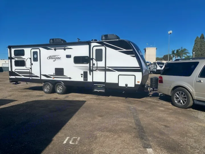 A bright white 'Imagine' travel trailer, an ideal rv rental, hitched to a tan SUV on a sunny outdoor lot, ready for departure.. Grand Design Imagine 2021