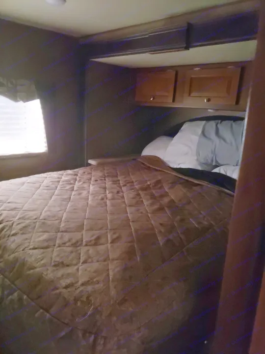 Compact RV rental bedroom featuring a brown quilted bed, storage cabinets, and window with blinds.. Coachman Leprechaun 2017