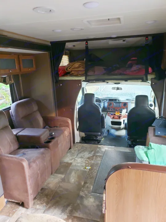 RV rental interior view: brown recliners, front cab, overhead bunk, and patterned floor. Ready for travel.. Coachman Leprechaun 2017