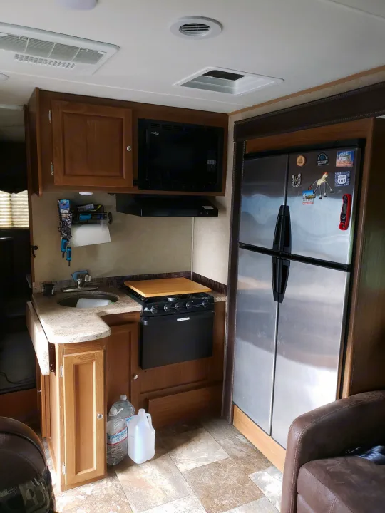 Inside an RV rental, a well-equipped kitchen features a large stainless steel refrigerator, microwave, sink, and stovetop.. Coachman Leprechaun 2017