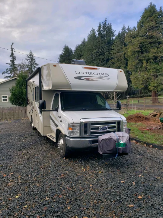 A white Coachmen Leprechaun Class C RV rental is parked on a gravel driveway. A gray covered item is on its front bumper.. Coachman Leprechaun 2017