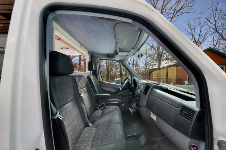 Experience this cozy rv rental's front cab, featuring dark gray seats, a functional dashboard, and a clear view of the winter scenery.. Mercedez Sprinter 2015