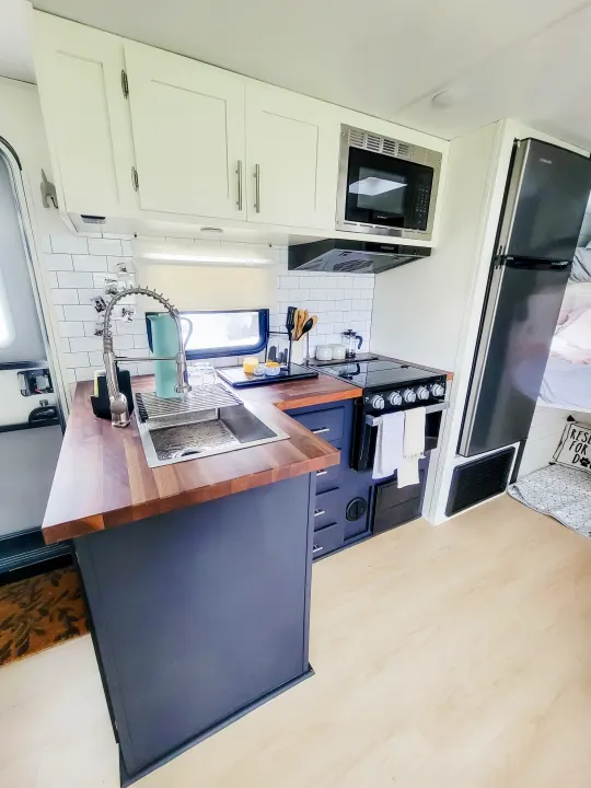 Inside a modern RV rental, a compact kitchen with white subway tile, sleek white and grey cabinets, wood counters, and black appliances.. Forest River Tracer 2022