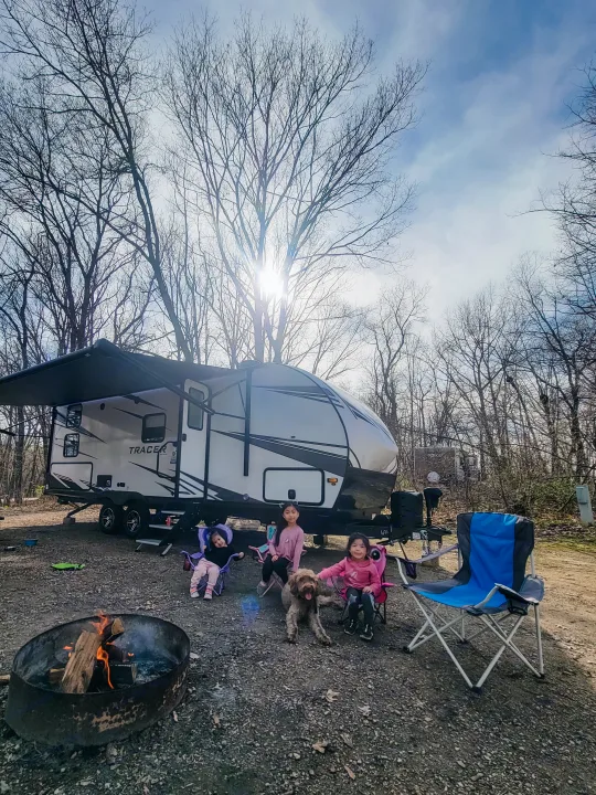 Family camping with their RV rental, dog, and campfire. Kids relax at the sunny campsite.. Forest River Tracer 2022