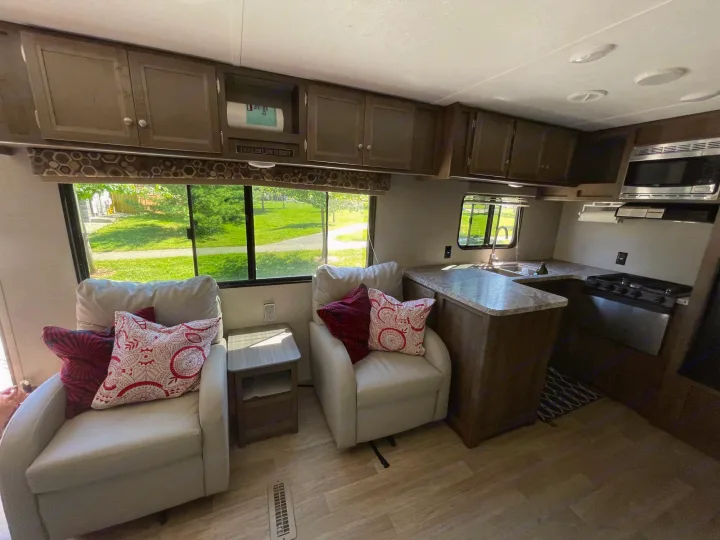 Inside this RV rental: Two comfy armchairs with pillows, a side table, and a compact kitchen area with stove and microwave.. Coleman Lantern 28.6 RK 2018