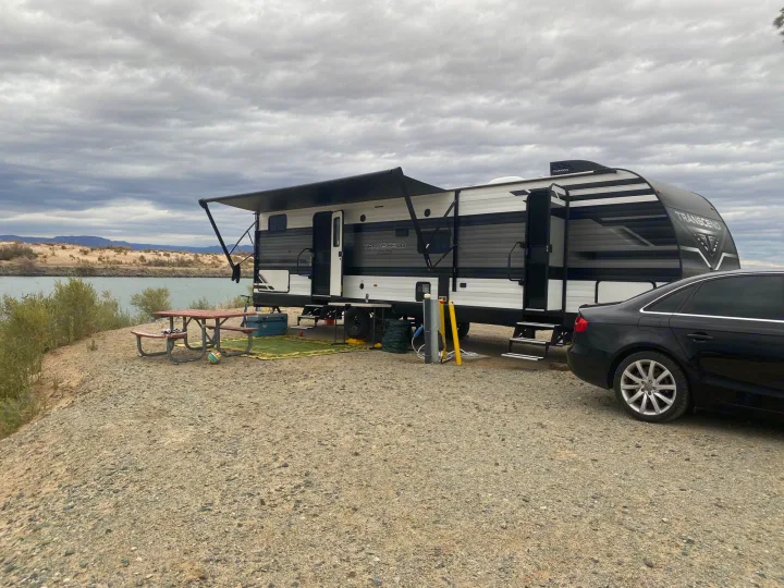 Spacious RV rental parked by a river, featuring an extended awning, picnic table, and utility hookups on a cloudy day.. Grand Design Transcend 2022