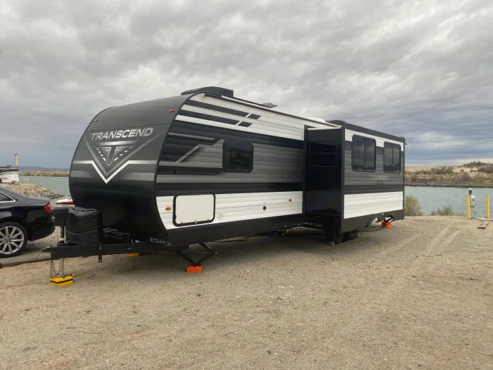 Ready for your next adventure? This Transcend travel trailer, an ideal RV rental, is set up by the water under a cloudy sky.. Grand Design Transcend 2022