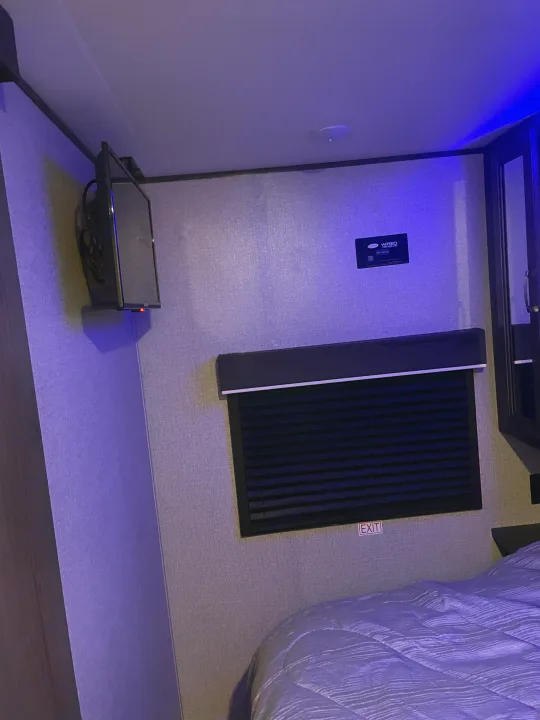 Compact RV rental bedroom featuring a wall-mounted TV, solar control panel, and window with dark blind. Blue LED lighting illuminates the bed.. Jayco Jay Flight 2021