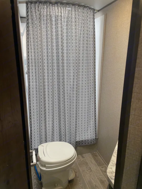 Compact RV rental bathroom with white toilet, wood-look floor, and a gray hexagonal patterned shower curtain.. Jayco Jay Flight 2021