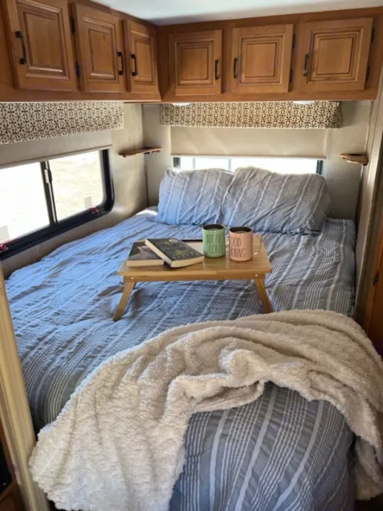 Cozy RV rental bedroom with blue striped bedding, a fluffy white blanket, and a bed tray with coffee mugs and a book.. Forest River Sunseeker 2016