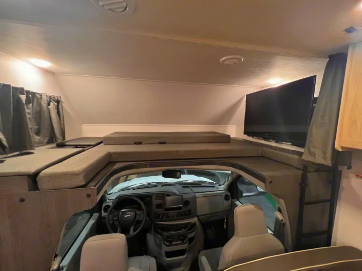 Interior of an RV rental: view of the cab-over bunk bed with TV, privacy curtains, and the driver's cockpit below.. Forest River Sunseeker 2024
