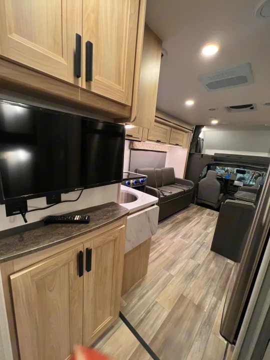 Inside a clean **rv rental**, showcasing a galley kitchen, flat-screen TV, and lounge seating with wood-look flooring.. Forest River Sunseeker 2024