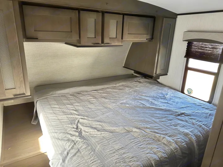 View inside an RV rental bedroom showing a gray bed, overhead cabinets, and a window with blinds.. Cruiser RV Embrace Ultra Lite 2022